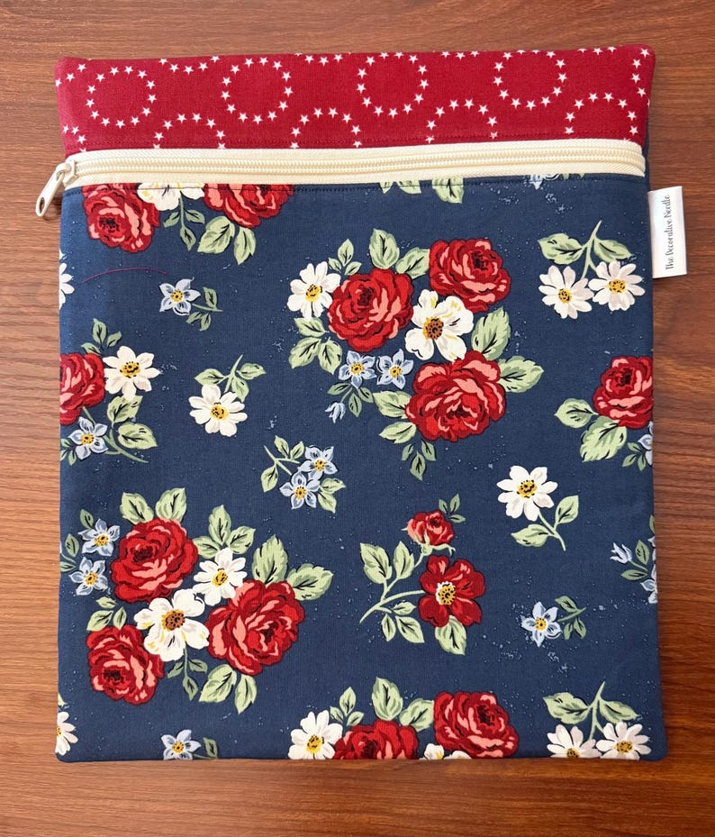 K&ouml;nnte beinhalten: Rechteckige Stofftasche mit Rei&szlig;verschluss. Der Hauptteil zeigt ein marineblaues Blumenmuster mit roten Rosen, wei&szlig;en G&auml;nsebl&uuml;mchen und gr&uuml;nen Bl&auml;ttern. Der obere Teil ist rot mit einem wei&szlig;en Sternmuster. Ein kleines wei&szlig;es Etikett ist angebracht.