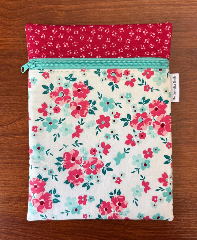 Puede incluir: Una bolsa con estampado floral y cremallera turquesa. La bolsa presenta un fondo blanco con flores rosas y turquesas. La secci&oacute;n superior es roja lisa con un estampado floral blanco. El texto "the decorative needle" est&aacute; impreso en el lateral.