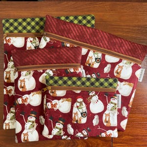 May include: A set of three fabric zipper pouches in varying sizes, featuring a dark red background with a repeating pattern of snowmen, foxes, and hedgehogs. The pouches have brown zippers and top bands in either dark red stripes or green plaid.