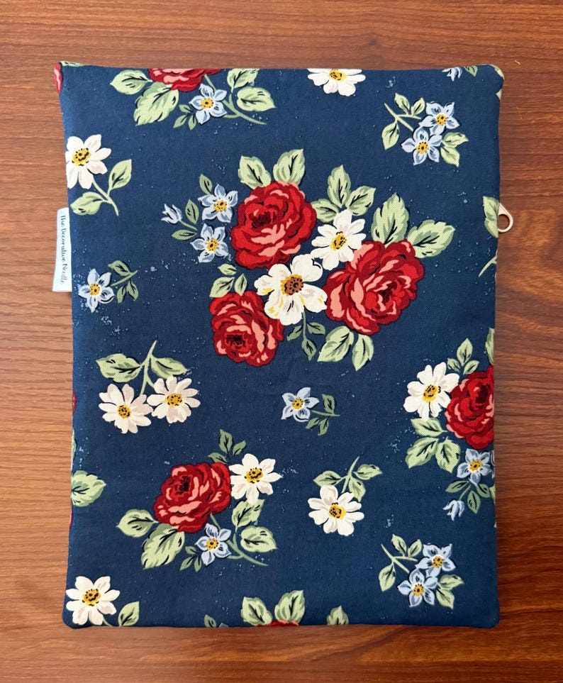 K&ouml;nnte beinhalten: Eine marineblaue Buchh&uuml;lle mit einem Blumenmuster aus roten Rosen, wei&szlig;en G&auml;nsebl&uuml;mchen und kleinen blauen Blumen mit gr&uuml;nen Bl&auml;ttern. Die H&uuml;lle hat ein kleines wei&szlig;es Etikett mit Text und einen Metallring.