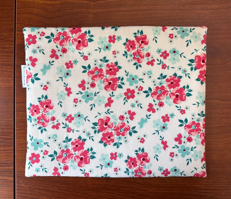 Puede incluir: Una funda rectangular con estampado floral. Presenta un fondo blanco con un patr&oacute;n repetido de flores rosas y turquesas y hojas verdes. La funda tiene un aspecto suave de tela y una peque&ntilde;a etiqueta blanca en el lateral.