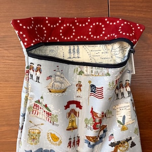 Op de afbeelding: Een lichtblauwe stoffen tas met ritssluiting, met een patriottische print van historische Amerikaanse beelden. De binnenvoering van de tas toont een kaartontwerp. De bovenrand is rood met witte sterpatronen.