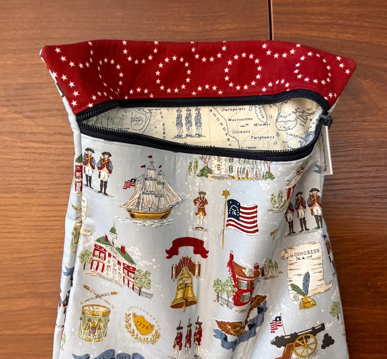 Op de afbeelding: Een lichtblauwe stoffen tas met ritssluiting, met een patriottische print van historische Amerikaanse beelden. De binnenvoering van de tas toont een kaartontwerp. De bovenrand is rood met witte sterpatronen.