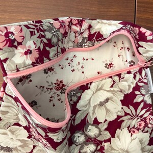 May include: A fabric pouch featuring a deep burgundy background with a large-scale floral pattern in shades of cream, light pink, and grey. The interior is lined with a white fabric featuring a small-scale floral print and a light pink zipper.