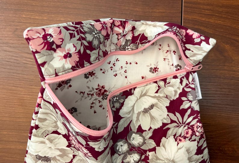 May include: A fabric pouch featuring a deep burgundy background with a large-scale floral pattern in shades of cream, light pink, and grey. The interior is lined with a white fabric featuring a small-scale floral print and a light pink zipper.