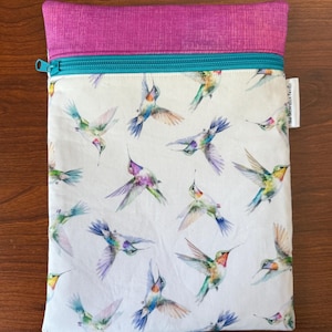 May include: A rectangular pouch with a white background, featuring a pattern of colorful hummingbirds in flight. The top is a textured magenta fabric, with a teal zipper and trim. A small white tag is on the right side.