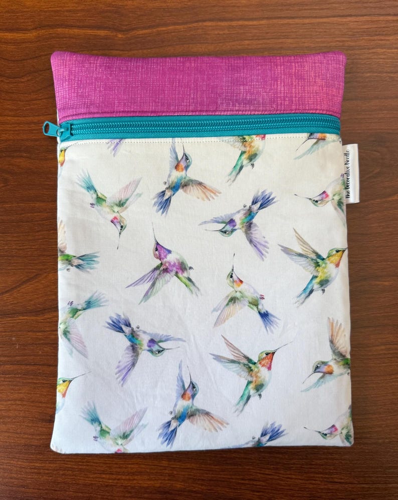 May include: A rectangular pouch with a white background, featuring a pattern of colorful hummingbirds in flight. The top is a textured magenta fabric, with a teal zipper and trim. A small white tag is on the right side.