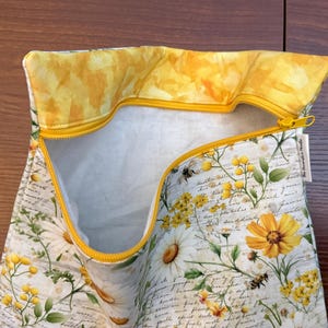K&ouml;nnte beinhalten: Ein Stoffbeutel mit Rei&szlig;verschluss auf wei&szlig;em Grund, bedruckt mit einem botanischen Muster aus gelben und wei&szlig;en Blumen, gr&uuml;nen Bl&auml;ttern und Bienen &uuml;ber einer handschriftlichen Textoptik. Das Innenfutter ist gelb und der Rei&szlig;verschluss ist leuchtend gelb.