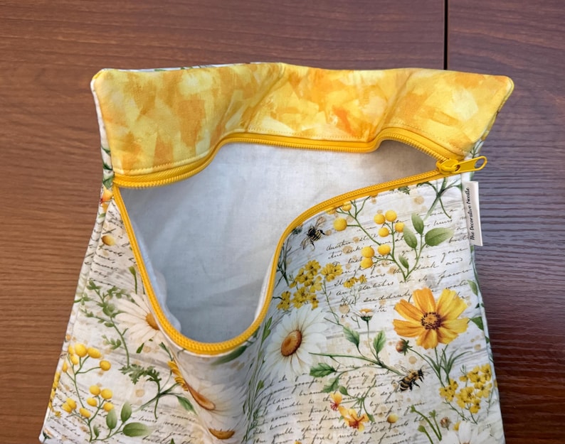 K&ouml;nnte beinhalten: Ein Stoffbeutel mit Rei&szlig;verschluss auf wei&szlig;em Grund, bedruckt mit einem botanischen Muster aus gelben und wei&szlig;en Blumen, gr&uuml;nen Bl&auml;ttern und Bienen &uuml;ber einer handschriftlichen Textoptik. Das Innenfutter ist gelb und der Rei&szlig;verschluss ist leuchtend gelb.