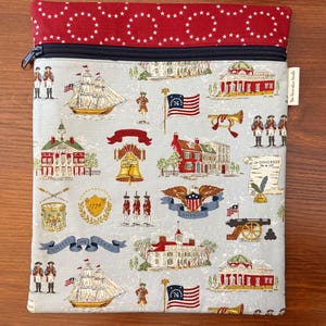 Op de afbeelding: Een etui met ritssluiting met een patriottisch thema. De hoofdbody heeft een lichtblauwe achtergrond met herhaalde illustraties van historische Amerikaanse bezienswaardigheden, vlaggen en soldaten. Het bovenste gedeelte is rood met witte sterpatronen.