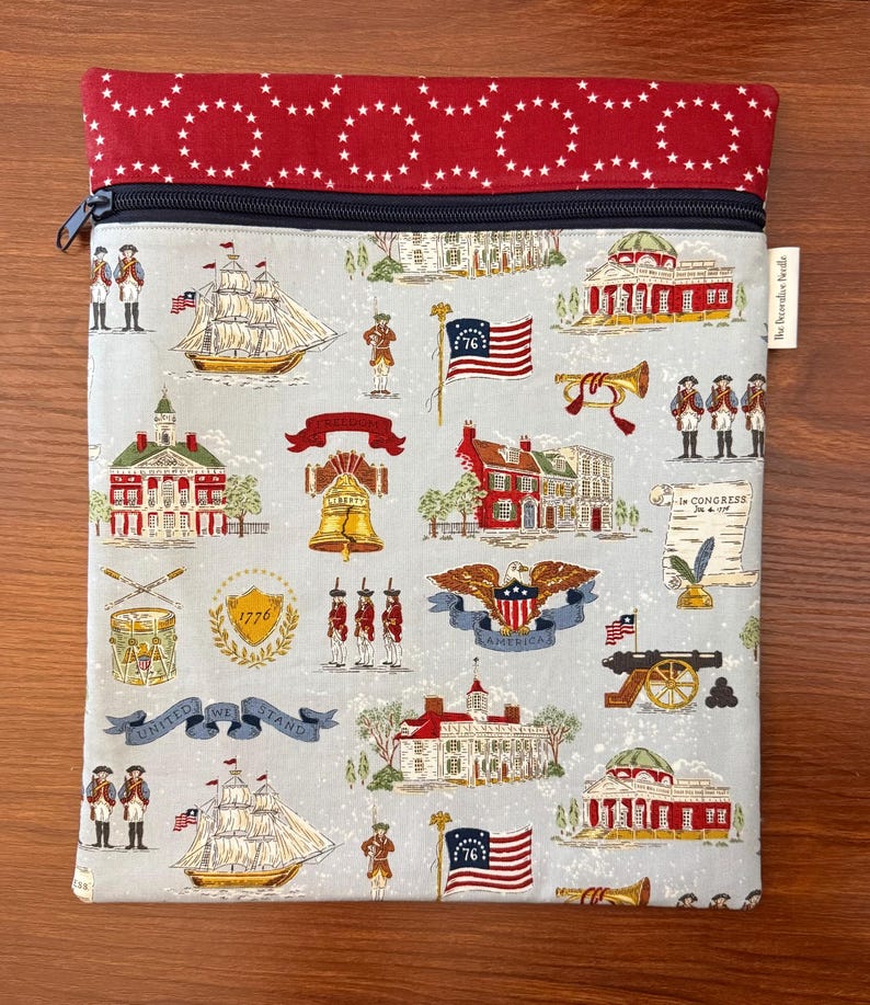 Op de afbeelding: Een etui met ritssluiting met een patriottisch thema. De hoofdbody heeft een lichtblauwe achtergrond met herhaalde illustraties van historische Amerikaanse bezienswaardigheden, vlaggen en soldaten. Het bovenste gedeelte is rood met witte sterpatronen.