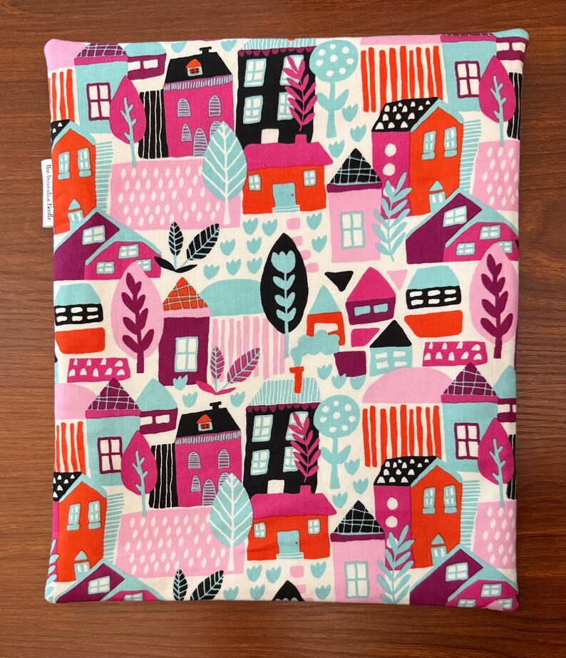 Puede incluir: Una bolsa de tela rectangular con un patr&oacute;n repetitivo y colorido de casas, &aacute;rboles y formas abstractas en tonos rosa, naranja, azul claro, morado, negro y blanco. Una peque&ntilde;a etiqueta de tela blanca con texto negro est&aacute; unida al lado izquierdo.