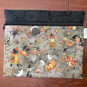 May include: A rectangular zippered pouch with a Halloween-themed pattern. The design features witches, pumpkins, black cats, ghosts, and stars on a gray background. The top section is black with a star and circle pattern.