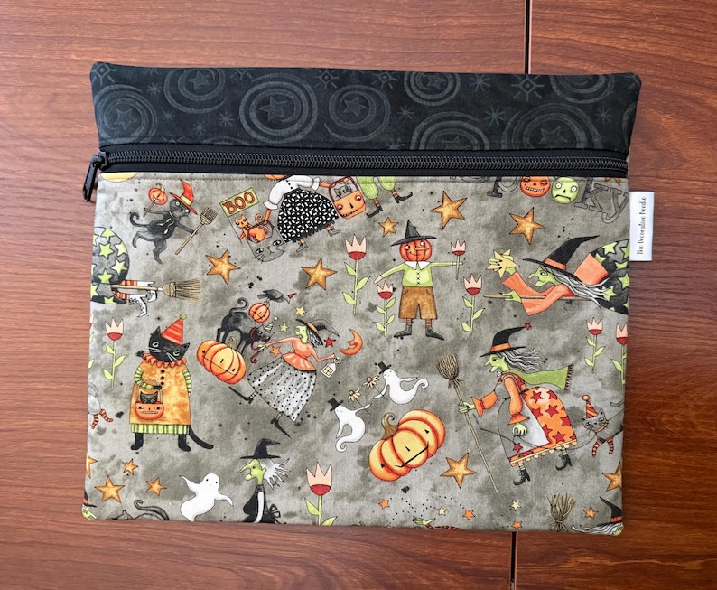 May include: A rectangular zippered pouch with a Halloween-themed pattern. The design features witches, pumpkins, black cats, ghosts, and stars on a gray background. The top section is black with a star and circle pattern.