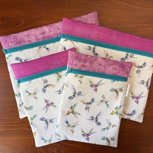 May include: Four zippered pouches with a white background and a hummingbird pattern. The pouches have a purple top, teal zipper, and a purple patterned trim. The pouches are made of fabric.