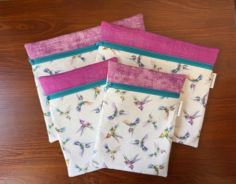 May include: Four zippered pouches with a white background and a hummingbird pattern. The pouches have a purple top, teal zipper, and a purple patterned trim. The pouches are made of fabric.