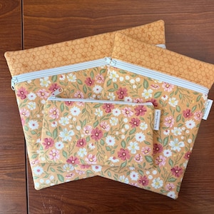 May include: Three zippered pouches in varying sizes. The pouches feature a floral pattern with pink, white, and green flowers on a yellow background. The top border is a honeycomb pattern. The zippers are silver with a light blue stripe.