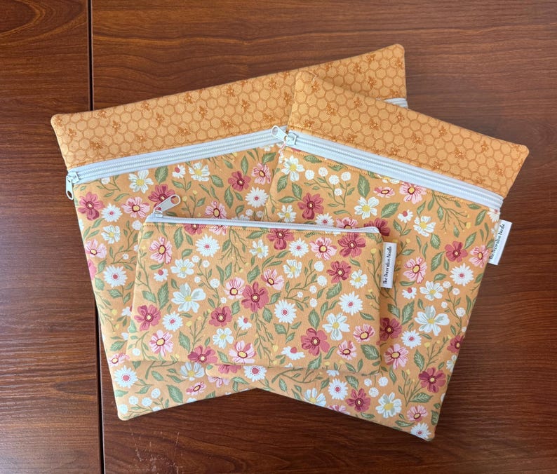 May include: Three zippered pouches in varying sizes. The pouches feature a floral pattern with pink, white, and green flowers on a yellow background. The top border is a honeycomb pattern. The zippers are silver with a light blue stripe.