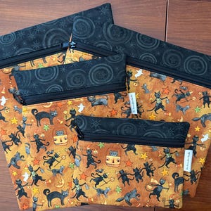 May include: A set of four zippered pouches in varying sizes. The pouches feature a Halloween-themed pattern with black cats, stars, and ghosts on an orange background. The top and zipper are black.