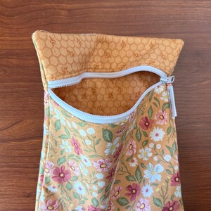 May include: A fabric pouch with a white zipper, featuring a floral pattern with pink, white, and green flowers on a yellow background. The top section has a honeycomb pattern. The pouch is open, revealing the interior.