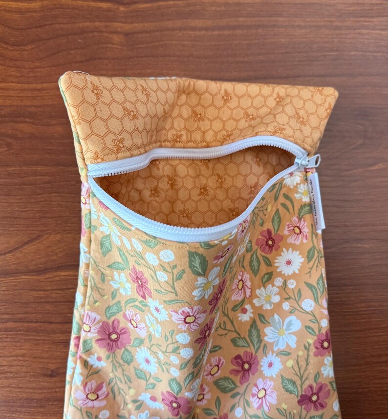May include: A fabric pouch with a white zipper, featuring a floral pattern with pink, white, and green flowers on a yellow background. The top section has a honeycomb pattern. The pouch is open, revealing the interior.