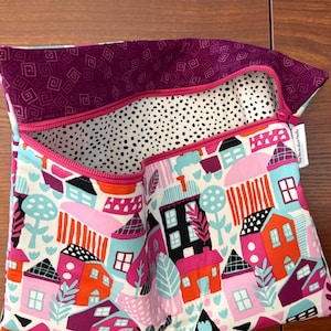 Puede incluir: Un estuche de tela con cremallera que presenta un patr&oacute;n colorido de casas y &aacute;rboles en tonos rosa, naranja, azul y negro sobre un fondo blanco. El interior est&aacute; forrado con una tela de lunares blancos y negros, y el borde es morado.