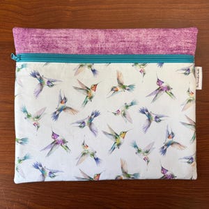 May include: A rectangular zippered pouch with a white background, covered in watercolor hummingbirds in flight. The top section is a textured purple fabric, and the zipper is teal. The label reads "The Decorative Needle".