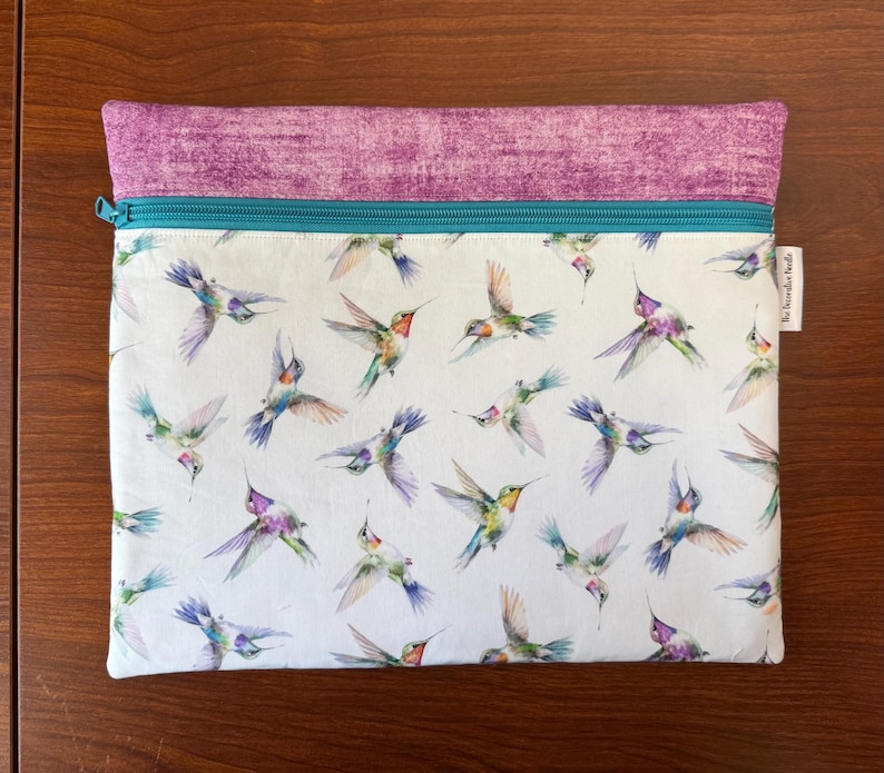 May include: A rectangular zippered pouch with a white background, covered in watercolor hummingbirds in flight. The top section is a textured purple fabric, and the zipper is teal. The label reads "The Decorative Needle".