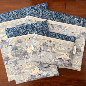 May include: A set of four zippered pouches in varying sizes, featuring a light blue coastal town print with sailboats, lighthouses, and houses. Each pouch has a dark blue top band with a white botanical pattern and a white zipper closure.
