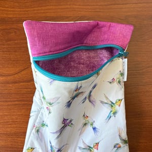 May include: A white zippered pouch with a vibrant hummingbird print. The pouch features a teal zipper, a magenta interior, and a magenta textured top. The words "the interactive tweedle" are printed on a small white tag.