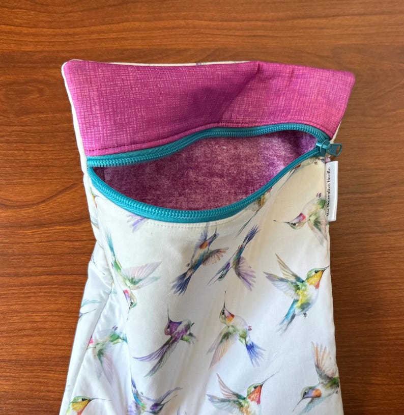 May include: A white zippered pouch with a vibrant hummingbird print. The pouch features a teal zipper, a magenta interior, and a magenta textured top. The words "the interactive tweedle" are printed on a small white tag.