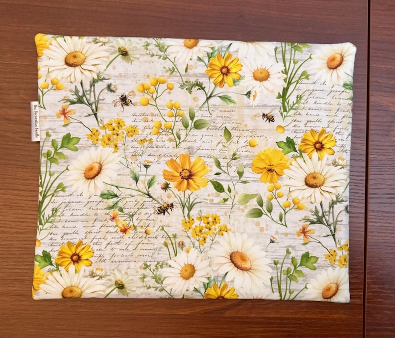 K&ouml;nnte beinhalten: Ein quadratischer Stoffartikel mit einem Muster aus wei&szlig;en G&auml;nsebl&uuml;mchen, gelben Wildblumen und kleinen Bienen auf einem Hintergrund aus hellgrauem Schreibschrifttext. Der Stoff hat ein kleines wei&szlig;es Etikett mit dem Text The Decoration Needle.