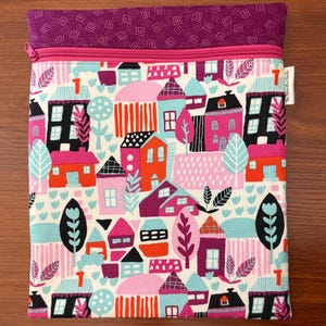 Puede incluir: Una bolsa de tela rectangular con cierre de cremallera y una banda superior de tela morada con un patr&oacute;n geom&eacute;trico. El cuerpo principal muestra un estampado colorido de casas y &aacute;rboles estilizados en tonos rosa, naranja, negro y azul claro.