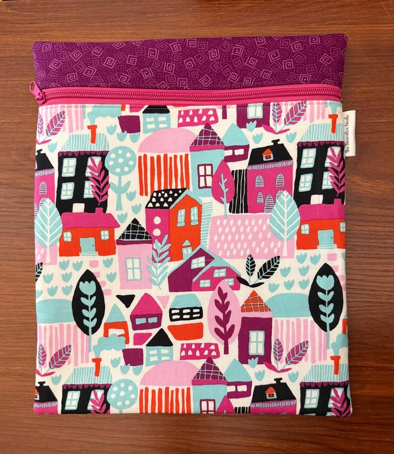 Puede incluir: Una bolsa de tela rectangular con cierre de cremallera y una banda superior de tela morada con un patr&oacute;n geom&eacute;trico. El cuerpo principal muestra un estampado colorido de casas y &aacute;rboles estilizados en tonos rosa, naranja, negro y azul claro.