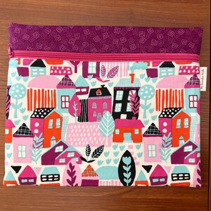 Puede incluir: Un estuche de tela rectangular con una secci&oacute;n superior de tela morada con un patr&oacute;n geom&eacute;trico blanco y un cuerpo principal con un estampado colorido de casas, &aacute;rboles y follaje en tonos rosa, naranja, turquesa y negro, cerrado con una cremallera rosa.