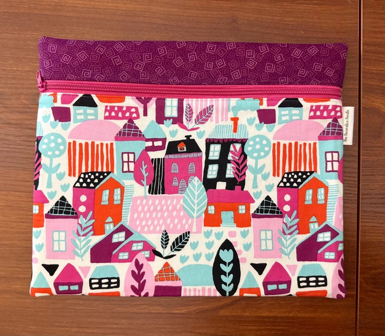 Puede incluir: Un estuche de tela rectangular con una secci&oacute;n superior de tela morada con un patr&oacute;n geom&eacute;trico blanco y un cuerpo principal con un estampado colorido de casas, &aacute;rboles y follaje en tonos rosa, naranja, turquesa y negro, cerrado con una cremallera rosa.