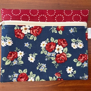 K&ouml;nnte beinhalten: Rechteckige Rei&szlig;verschlusstasche mit floralem Muster. Der Hauptteil ist marineblau mit roten Rosen, wei&szlig;en G&auml;nsebl&uuml;mchen und kleinen blauen Blumen. Die obere Klappe ist rot mit einem wei&szlig;en Sternmuster. Der Rei&szlig;verschluss ist beige.