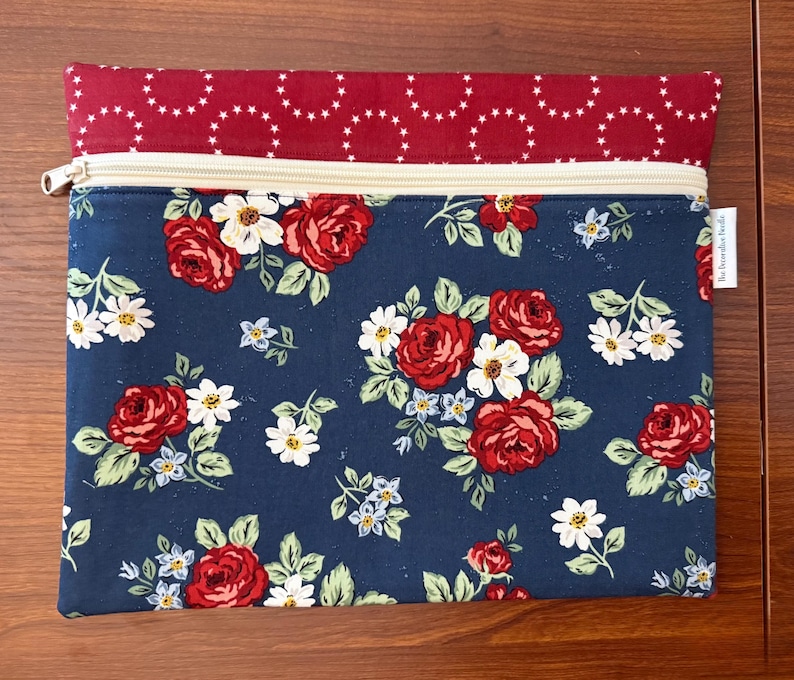 K&ouml;nnte beinhalten: Rechteckige Rei&szlig;verschlusstasche mit floralem Muster. Der Hauptteil ist marineblau mit roten Rosen, wei&szlig;en G&auml;nsebl&uuml;mchen und kleinen blauen Blumen. Die obere Klappe ist rot mit einem wei&szlig;en Sternmuster. Der Rei&szlig;verschluss ist beige.