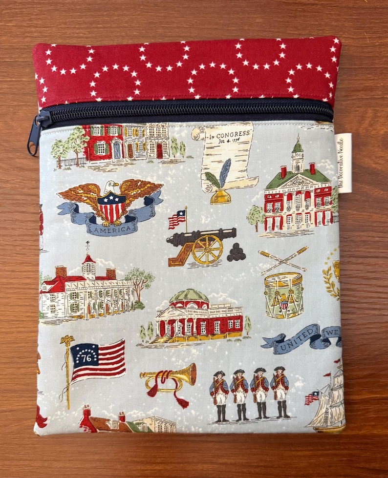 Op de afbeelding: Een rechthoekig etui met ritssluiting. De bovenkant is rood met witte sterren en de body is lichtblauw met patriottische illustraties. Deze omvatten een adelaar, gebouwen, vlaggen en soldaten. De rits is marineblauw.