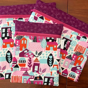 Puede incluir: Tres estuches rectangulares de tela con un patr&oacute;n colorido de casas, &aacute;rboles y follaje en tonos rosa, naranja, turquesa y negro. Cada estuche tiene una banda superior de color morado oscuro con un patr&oacute;n geom&eacute;trico y una cremallera de color rosa brillante.