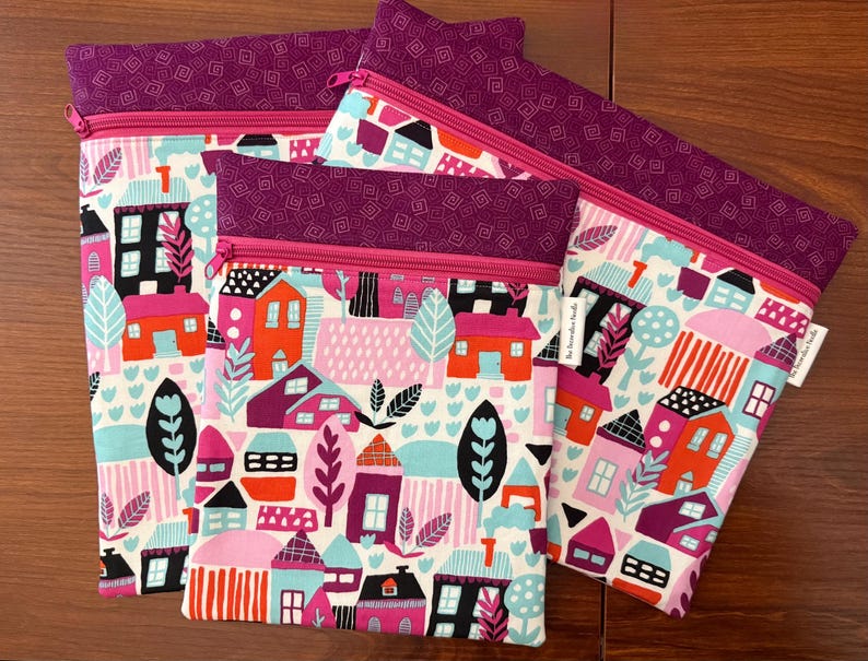 Puede incluir: Tres estuches rectangulares de tela con un patr&oacute;n colorido de casas, &aacute;rboles y follaje en tonos rosa, naranja, turquesa y negro. Cada estuche tiene una banda superior de color morado oscuro con un patr&oacute;n geom&eacute;trico y una cremallera de color rosa brillante.