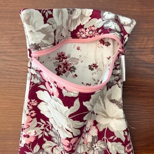 May include: A zippered pouch featuring a floral pattern with deep burgundy, light pink, and cream-colored blossoms. The interior lining is cream with a smaller floral print. A light pink zipper secures the opening, and a small white fabric tag is attached.