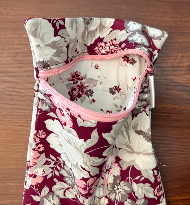 May include: A zippered pouch featuring a floral pattern with deep burgundy, light pink, and cream-colored blossoms. The interior lining is cream with a smaller floral print. A light pink zipper secures the opening, and a small white fabric tag is attached.