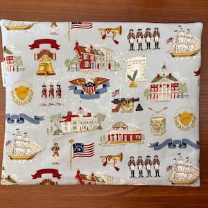 Op de afbeelding: Een lichtblauw stoffen item met een herhalend patroon van Amerikaanse historische beelden. Het ontwerp bevat de Liberty Bell, vlaggen, schepen, soldaten en gebouwen. De woorden "1776", "America" en "United We Stand" zijn ook zichtbaar.