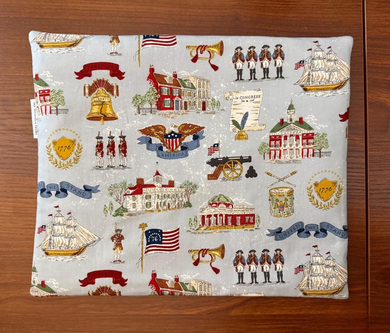 Op de afbeelding: Een lichtblauw stoffen item met een herhalend patroon van Amerikaanse historische beelden. Het ontwerp bevat de Liberty Bell, vlaggen, schepen, soldaten en gebouwen. De woorden "1776", "America" en "United We Stand" zijn ook zichtbaar.