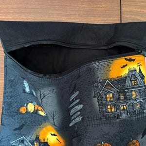 May include: A black zippered pouch featuring a Halloween-themed print with a haunted house, glowing orange moon, pumpkins, bats, and a signpost. The interior is lined with black fabric, and a small white tag labeled The Decorative Needle is attached.