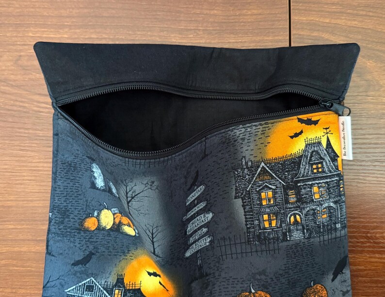 May include: A black zippered pouch featuring a Halloween-themed print with a haunted house, glowing orange moon, pumpkins, bats, and a signpost. The interior is lined with black fabric, and a small white tag labeled The Decorative Needle is attached.
