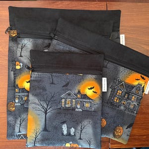 May include: Three rectangular zippered pouches of varying sizes, each featuring a black fabric top and a gray patterned lower section. The print displays a spooky scene with haunted houses, bare trees, orange moons, bats, and jack-o'-lanterns.