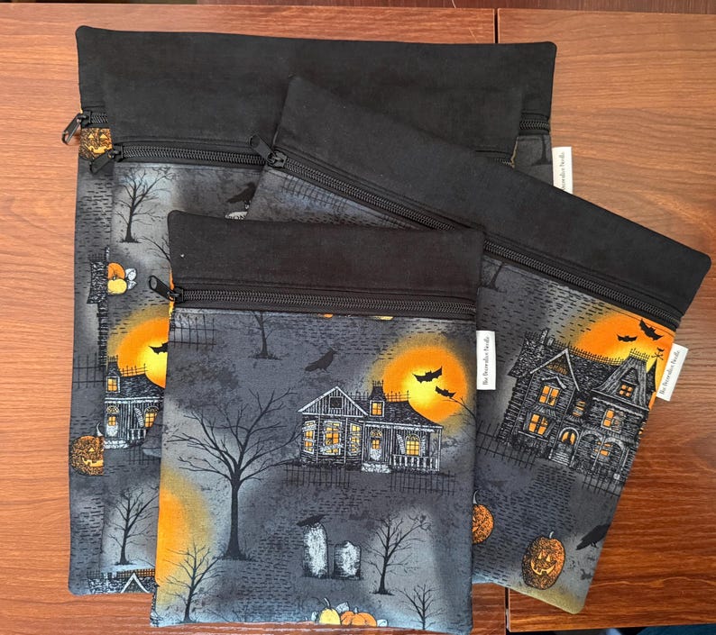 May include: Three rectangular zippered pouches of varying sizes, each featuring a black fabric top and a gray patterned lower section. The print displays a spooky scene with haunted houses, bare trees, orange moons, bats, and jack-o'-lanterns.