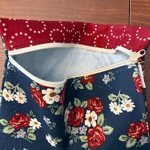 K&ouml;nnte beinhalten: Eine Rei&szlig;verschlusstasche mit floralem Muster. Der marineblaue Stoff ist mit roten Rosen und wei&szlig;en G&auml;nsebl&uuml;mchen verziert. Der obere Rand ist tiefrot mit wei&szlig;en Sternen. Der Rei&szlig;verschluss ist beige.